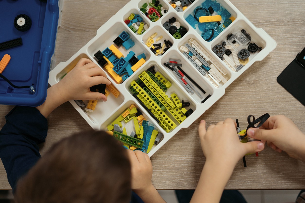 View of hands of incognito kid robot holding elements and working on project, details in box. Colorful pieces from building kit for children on table